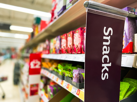 Close up color image depicting a sign with the word 'snacks' in a supermarket aisle. Focus on the sign with the snacks on the shelves defocused beyond. Room for copy space.