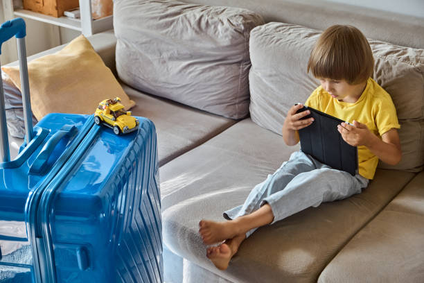 A sad child is sitting on a sofa using a tablet and waiting for departure. Family relocation, flight from the country