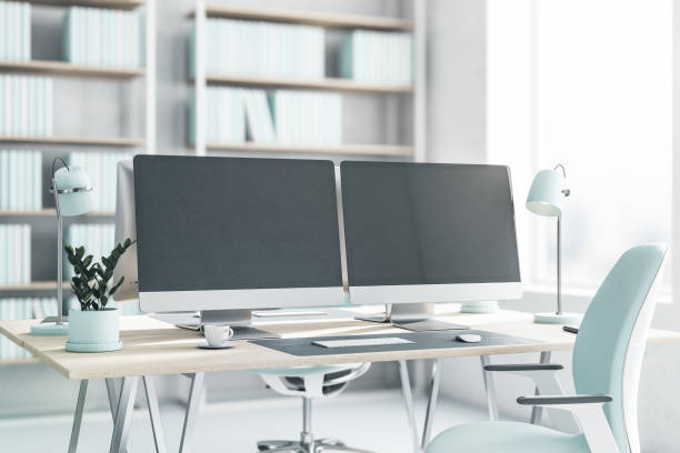 istock Side view on blank dark modern computer monitors on light wooden table with light blue lamp, flowerpot and chair on white floor in sunny office with big window. 3D rendering, mockup 1430388060