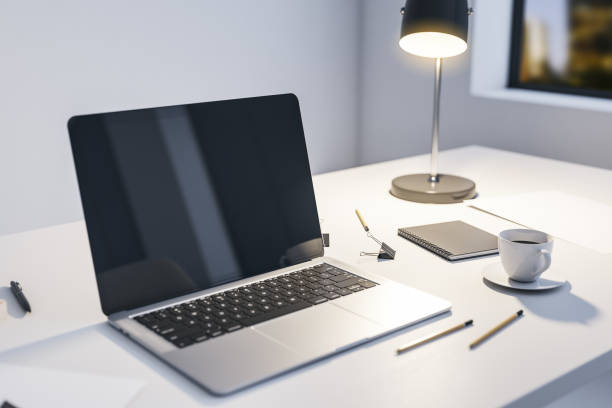 istock Perspective view on modern laptop blank dark screen on light table with illuminated lamp, office tools and coffee cup in light walls room and night city view from the window. 3D rendering, mockup 1430388232