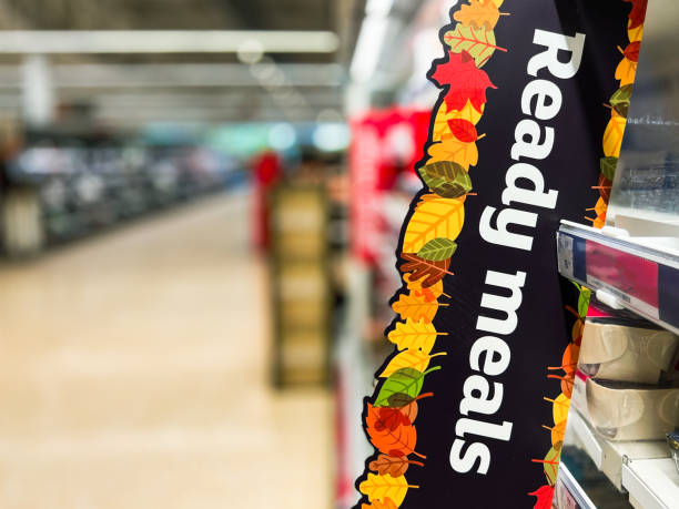 Close up color image depicting a sign with the words 'ready meals' in a supermarket aisle, with a selection of ready meals and tv dinners defocused on the shelves beyond.