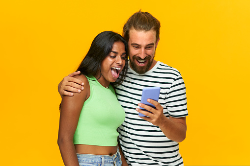 Image of multiracial couple surprised looking at mobile phone, isolated over yellow background, positive, excited people, man and woman screaming and looking at one cell, male showing to girlfriend.