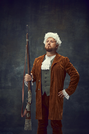 Hunter. Nobleman, medieval royal person in vintage hunting clothing with old gun isolated over dark background. Art, fashion, emotions, traditions, male hobby concept