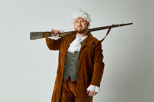 Serious young man in image of medieval person in vintage brown hunting suit and white wig with old hunting rifle isolated over grey background. Art, fashion, emotions, male hobby concept