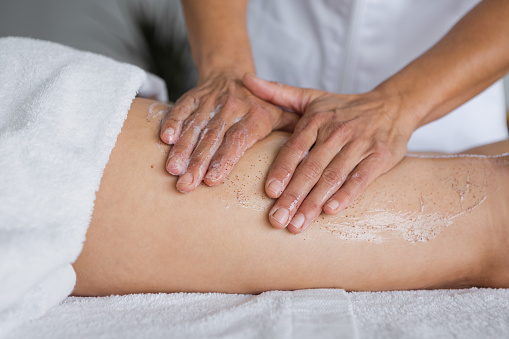Crop unrecognizable beautician rubbing scrub gel into leg skin of woman during anti cellulite massage session on table in parlor