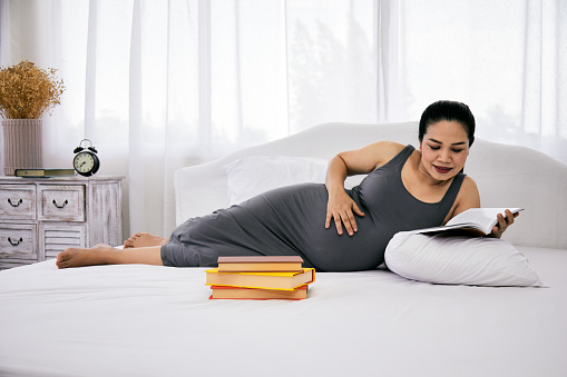 Asian Pregnant mother lying on white bed holding and reading book