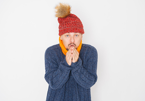 Young handsome man wearing in red hat and blue pullover feeling cold on white background. Gas crisis concept
