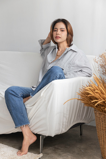 Smiling beautiful asian woman in gray shirt and blue jean stay at home sitting relax on cozy couch.