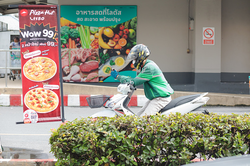 GRAB delivery person is waiting outside of supermarket in Bangkok. Scene is at a big Lotus supermarket in Ladprao. Man os wearing yellow crash helmet and is using mobile phone