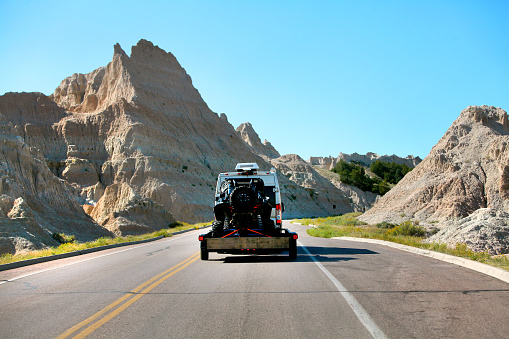 Traveling directly behind an sprinter adventure van pulling a trailer with a black off road vehicle between two ridges of the Badlands hills in South Dakota on a cloudless sunlit afternoon