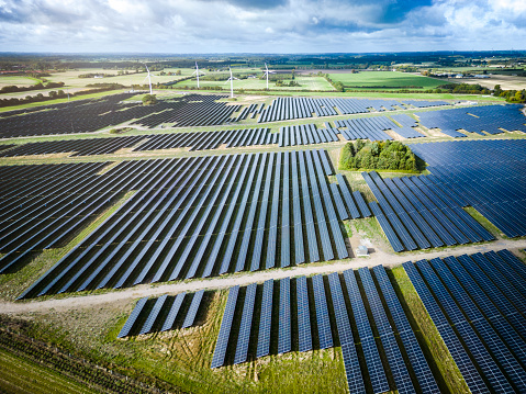 Sunlight on solar panel in a large solar energy farm in rural area in Denmark. Wind turbines are seen in the background. Aerial view shot with drone.
