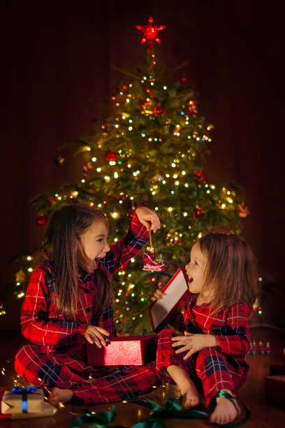 就学前の年齢の2人の面白い妹がクリスマスイブにプレゼントを開梱しています。子供たちはクリスマスツリーのおもちゃの入った箱を開けながら楽しそうに笑います。贈り物を受け取る幸福 - merrily ストックフォトと画像