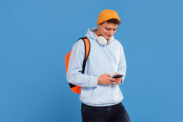istock Young fashionable student guy with cap and backpack smiling, using mobile phone, isolated over blue studio background 1430451741