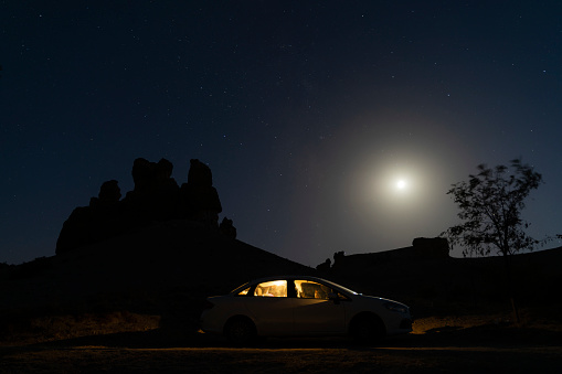 parked car in the countryside at dusk. The interior lights of the vehicle visible in the moonlight are on. fairy chimney and tree silhouette appear. Shot with a full frame camera.