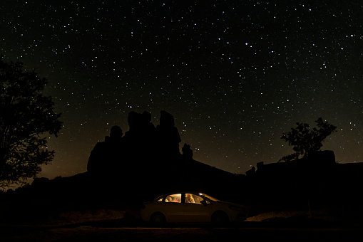 parked car in the countryside at dusk. A man is sitting in the vehicle. The interior lights of the vehicle visible in the moonlight are on. fairy chimney and tree silhouette appear. Shot with a full frame camera.