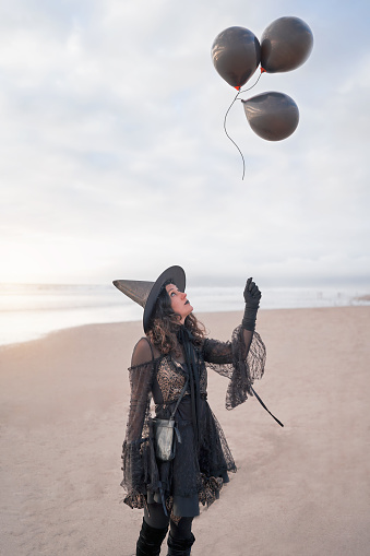 woman dressed as a witch standing on the beach releases black balloons to the sky