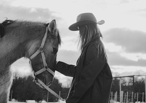 cowgirl petting her horse in the snow with hat and leather jacket on a cloudy day