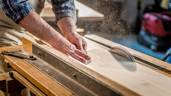 Detailed shot of woodwork, sawing material to proper size. Cropped view of carpenter's hands while using circular saw. Wood material processing in carpentry, handmade craftwork products.