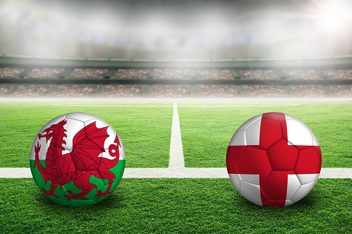 Wales vs England football in brightly lit outdoor stadium with painted English and Welsh flags. Focus on foreground and soccer ball with shallow depth of field on background and copy space.