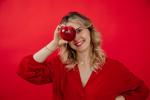 Young cheerful, merry, blissful blonde woman holding fresh apple near face, covering eye with smile on red isolated background. Healthy lifestyle and body care. Female health fertility, reproduction