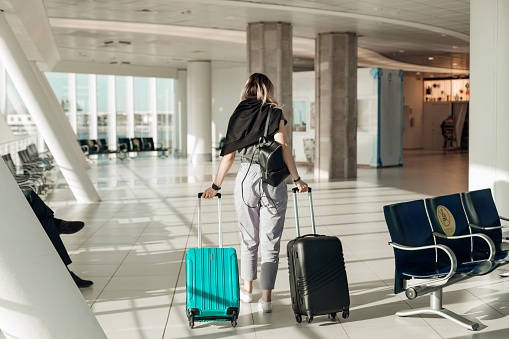 Passenger with baggage going to boarding plane in light airport with panoramic windows and rolling suitcases with clothing back view, airplane arriving. Young woman travel and relax on vacation.