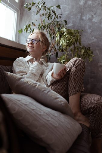 Vertical modern, smiling, pensive and cozy blond barefoot woman in glasses and warm clothes sit on sofa with cushions and drink hot tea, look out the window. Cold season and weekend relaxation at home