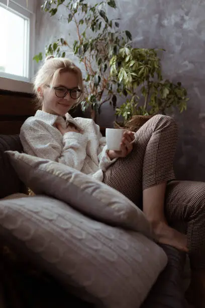 Vertical smiling, calm, glad blond woman in eyeglasses, in warm sweater sitting on soft sofa and drinking hot tea in houseplant arrangement. Day off and vacation, cozy weekend activity at home