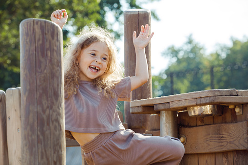 Summer children's games and fun outdoors. Happy child girl with blond gurly hair play on wooden playground on sunny day. Lifestyle and childhood concept.
