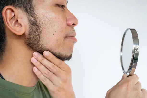 Photo of Cropped shot of Asian man touching his beard grows on a part of his lower face while looking his face by mirror.