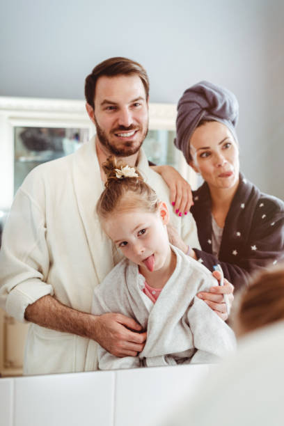 junge schöne familie in bademänteln, lächelnd und albernd vor einem badezimmerspiegel - mirror mother bathroom daughter stock-fotos und bilder