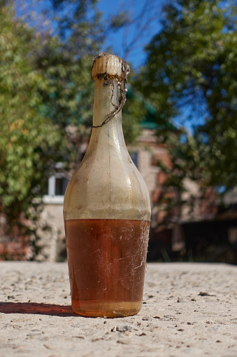 Old bottle of sunflower oil with sediment. Covered with cobwebs and dust, a bottle of brown thick liquid stands against the backdrop of trees, a blue sky and a village house. Selective focus.