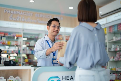 Asian mature man pharmacist explaining to young woman customer at pharmacy aisle.