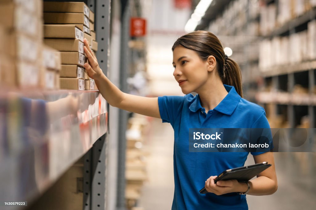 Young female worker in blue uniform checklist manage parcel box product in warehouse. Asian woman employee holding tablet working at store industry. Logistic import export concept. Warehouse Worker Stock Photo