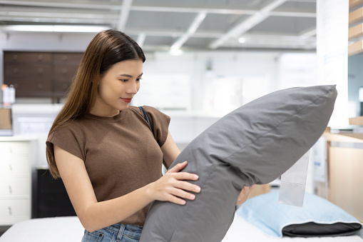 Young beautiful woman choosing pillow at retail furniture store. Housewife pick up the pillow and softness test buying for family. Female checking quality shopping at mattress store hall.