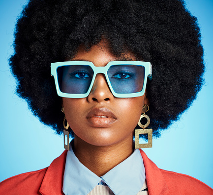 Fashion influencer and black woman sunglasses portrait with natural afro on blue studio wall. Trendy, cool and edgy african hipster girl with an assertive and serious face expression.