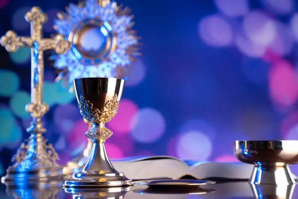 Catholic symbols composition. The Cross, monstrance,  Holy Bible and golden chalice on blue bokeh background.
