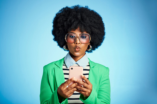Trendy, fashion and african girl model with an afro on a phone in a studio with a blue background. Young black hipster with edgy, funky and colorful clothes and sunglasses standing on a smartphone.
