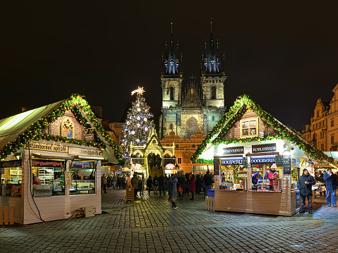 Prague, Czech Republic - December 6, 2017: Christmas market at the Old Town Square with the city's main Christmas tree in night. Two market stalls with food and drinks are located in the foreground. The Gothic church of Our Lady before Tyn is located on the background. Unknown people walk around the market stalls and stand at the lookout platform.