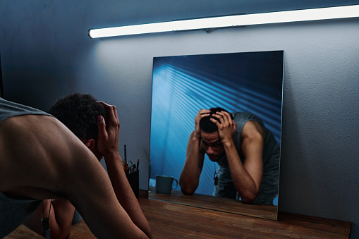 Young man with post traumatic disorder standing in front of mirror with his head in hands while feeling anxiety and helplessness