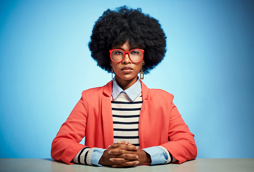 Serious, leader and confident black woman with afro while sitting at table against a blue studio background. Portrait of trendy, edgy attitude and stylish student or teacher female with glasses