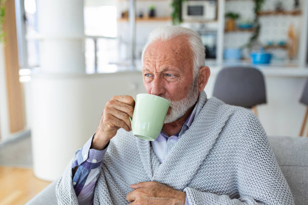 istock Senior man suffering from flu drinking tea while sitting wrapped in a blanket on the sofa at home. Sick older man with headache sitting under the blanket in the living room. Man with a cold lying on the sofa holding a mug of hot tea. 1430608842