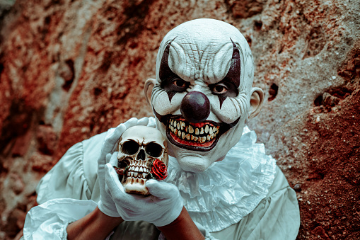 closeup of a creepy evil clown, wearing a gray costume with a white ruff, having a skull in his hands in front of the old and rusty exterior wall of an abandoned house