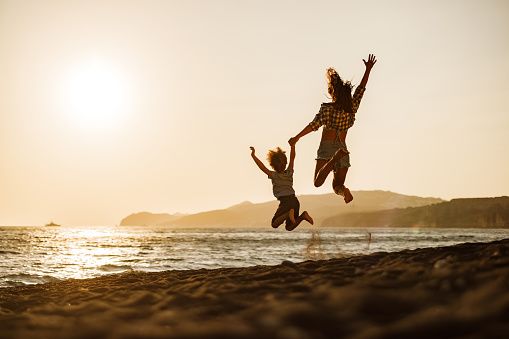 Rear view of single mother and son having fun while holding hands and jumping on the beach at sunset. Copy space.