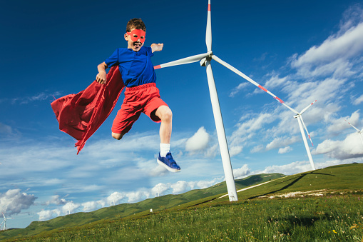 Superhero kid is in front of the wind farm. Little child plays superhero.