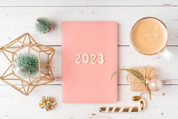 Christmas pink notepad with 2023 letters text. Flat lay on white wooden table background with planner, cup of coffee, candle, Christmas decoration, notebook and stationery. Top view Desktop.