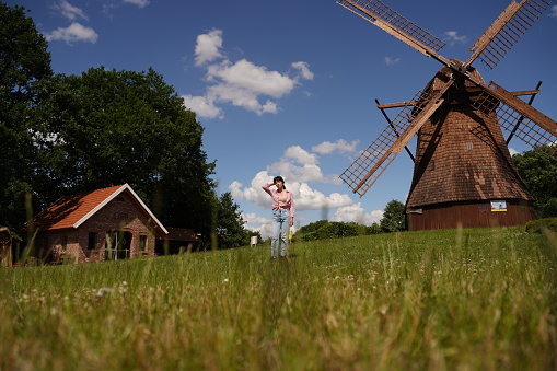 A young girl stands on a summer meadow against the background of old abandoned buildings. A woman with black hair dressed in a summer shirt and jeans, she threw her hand behind her head and looks at the clear blue sky with sparse white clouds. Selective focus.