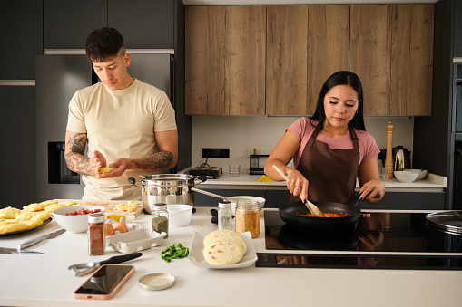 Two young hispanic people moulding llapingachos dough and cooking in a pan at kitchen.