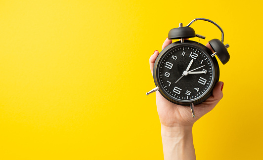 Black friday concept. First person top view photo of female hand holding black alarm clock on isolated yellow background with empty space