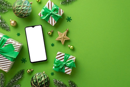 Christmas concept. Top view photo of smartphone gift boxes gold and green baubles star ornament confetti and pine branches in hoarfrost on isolated green background with blank space