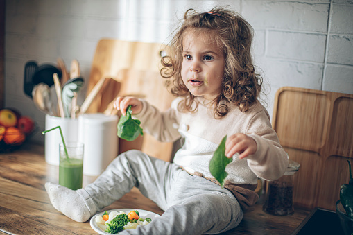 Cute baby girl eating green vegetables on a kitchen counter at home